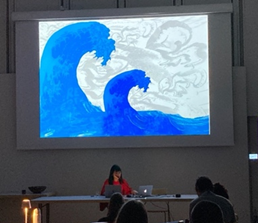 presenter sitting in front of a large image of two blue waves